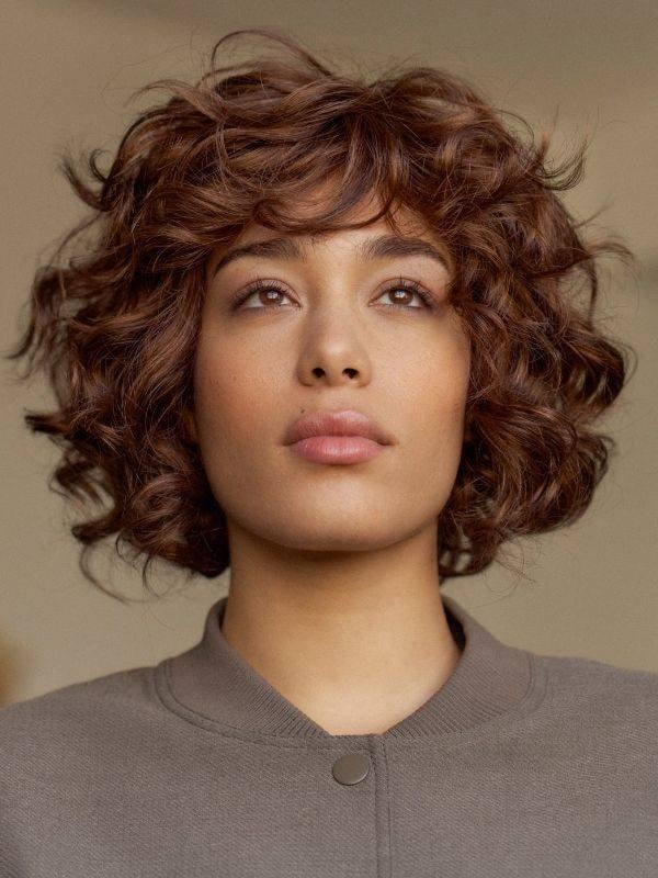 Perruque Kim NJ Création mi-longue bouclée, fibre synthétique effet naturel, frange volumineuse et confort médical antiglisse.